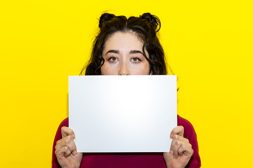 young woman holding and hiding her face on a poster. Girl holding a blank poster on isolated background