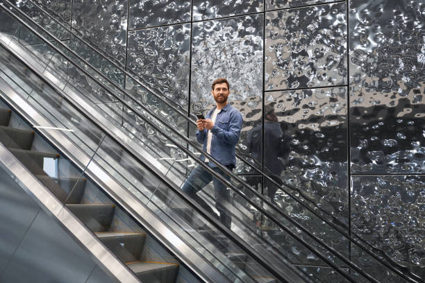 smiling bearded male with mobile phone, lifting up on escalator, while looking into distance. - escalator shopping mall shopping transparent imagens e fotografias de stock
