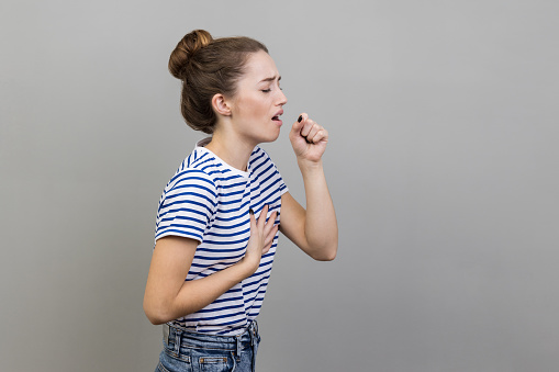 Side view portrait of sick unhealthy woman wearing striped T-shirt having cough and feeling pain in her lungs, catching cold, flu symptoms. Indoor studio shot isolated on gray background.