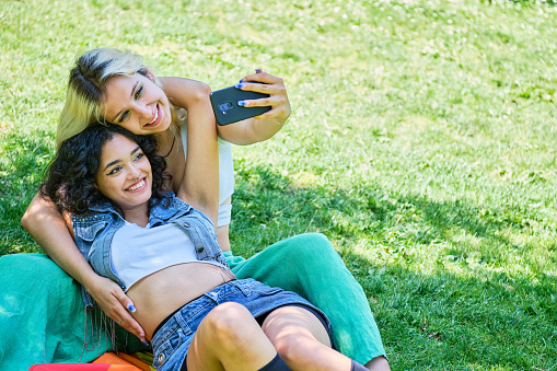 young lesbian couple smiling hugging sitting on the grass in a park taking a selfie