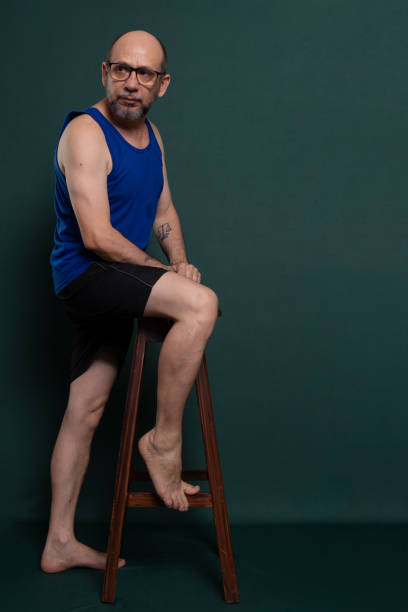 Bearded, bald white man leaning on a wooden stool posing for a photo.