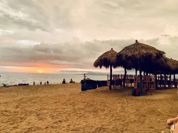 Tiki umbrellas and beach chairs watch the sun submerge into a tropical island horizon highlighting a cotton-candy sky among the overcast cloudiness.