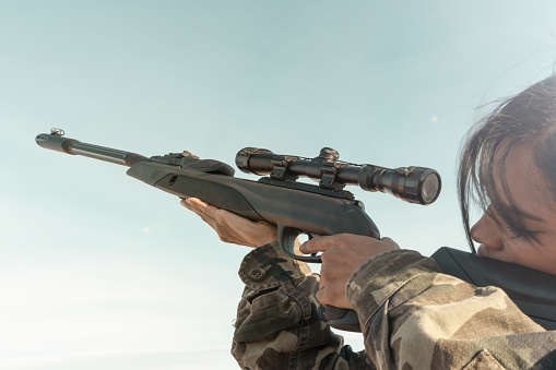 Close-up of a rifle with an optical sight in the hands of a black African woman in military uniform, aiming at the optical sight while. Military action, sniper shooting process.