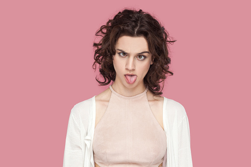 Portrait of crazy fool woman with curly hair wearing casual style outfit having foolish facial expression, grimacing, showing bad manners. Indoor studio shot isolated on pink background.