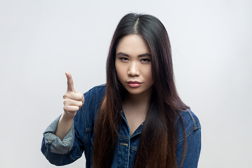 Portrait of serious beautiful brunette woman in blue denim jacket standing with raised finger, warning you, looking at camera with strict expression. Indoor studio shot isolated on gray background.