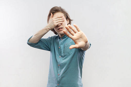 Stop, I don't want to see it. Portrait of young man standing, with stop gesture and covered his eyes, sees something shameful, wearing blue shirt. Indoor studio shot isolated on gray background.