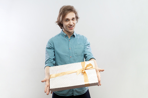 Portrait of satisfied friendly handsome young man standing with gift in hands, giving present to her girl friend on birthday, wearing blue shirt. Indoor studio shot isolated on gray background.