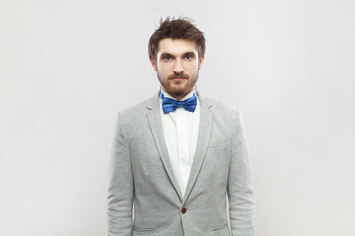 Portrait of calm positive bearded man standing looking at camera, having relaxed facial expression, wearing grey suit and blue bow tie. Indoor studio shot isolated on gray background.