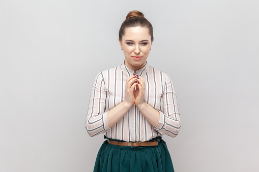 Portrait of cunning young adult woman wearing striped shirt and green skirt keeps hands together, having slander plan, pondering. Indoor studio shot isolated on gray background.