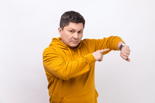 You are late. Portrait of serious bossy man standing, looking at camera and pointing at his smart watch, wearing urban style hoodie. Indoor studio shot isolated on white background.
