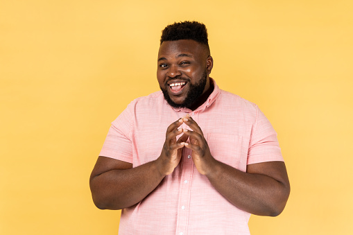 Portrait of cunning bearded man wearing pink shirt clasping hands and planning evil tricky prank or scheming, cheating with sly smile. Indoor studio shot isolated on yellow background.