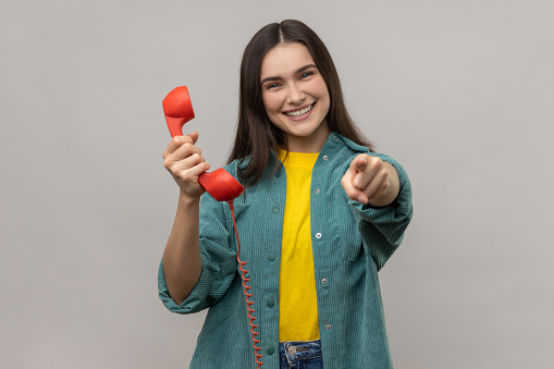 Attractive happy woman holding red landline phone and pointing to camera with finger, call me, I am waiting, wearing casual style jacket. Indoor studio shot isolated on gray background.