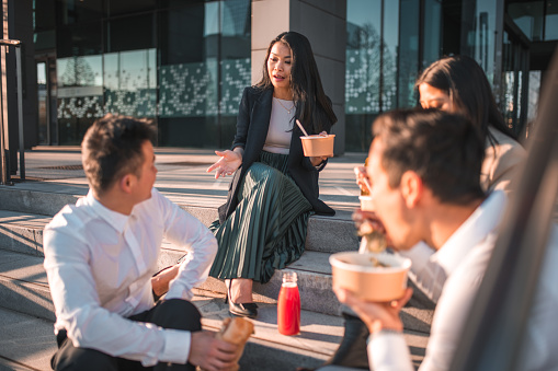 Embracing the lunchtime calm, Asian business professionals sit on the stairs outdoors, indulging in takeaway food bowls and sharing a peaceful moment.