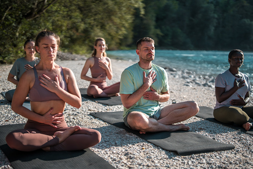 A varied group of people engages in breathing exercises, representing diverse backgrounds, on yoga mats by the tranquil river.