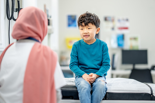 A young boy of Asian decent, sits up on an exam table during a routine check up.  His female Muslim doctor is seated in front of him as the two talk during the appointment.