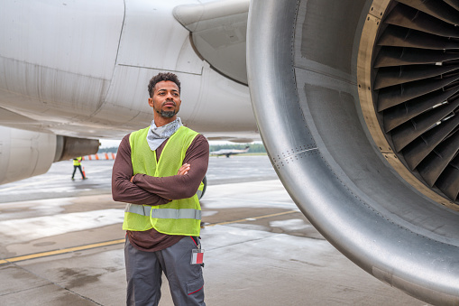 Three-quarter length shot of a Mixed Race male worker standing next to a plane turbine. He is with his arms crossed and looking away.