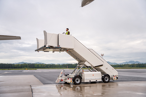 Mid adult male moving an airstair at the airport. He is working outdoors on a cloudy and rainy day.