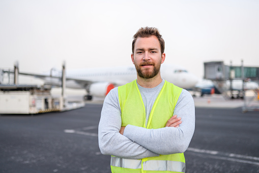 Portrait of a Caucasian aircraft maintenance technician with hands crossed. Standing at the airfield wearing a reflective vest.