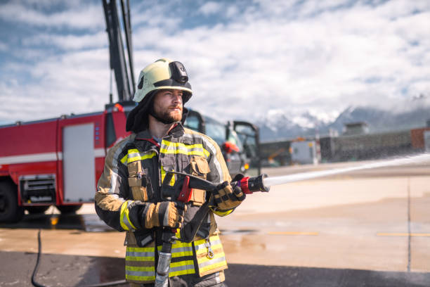 Young adult fireman using a water gun at the airport. Standing on a runway spraying water on a fire while wearing fire protection suit.