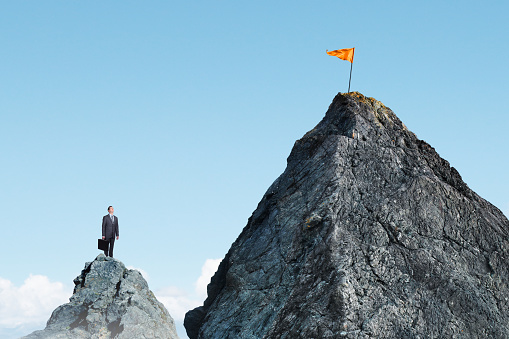 A businessman stands on a small mountaintop and looks up at an orange flag planted on top of a taller mountaintop.