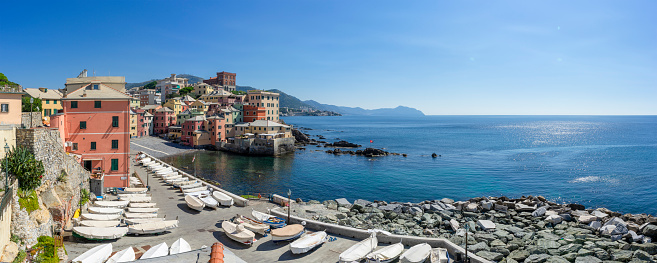 A panoramic view of a tranquil Mediterranean village by the sea, showcasing a vibrant tapestry of pastel-hued buildings cascading towards a cobalt blue ocean. White boats rest on the pebbled shore, awaiting their next voyage on the sparkling waters under the clear azure sky.