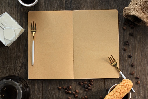 Blank Open Book for Cook Book/Recipe/Text with Cake Fork, Craquelin Eclair, Coffee Bean, Coffee Pot, and Milk. Concept for Menu Book on Brown Wooden Background