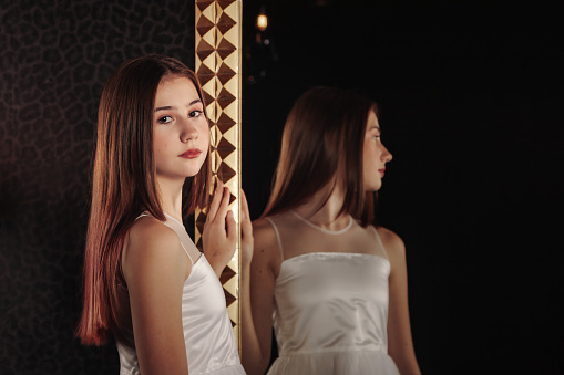 Pensive teenage lady in elegant white dress posing at large mirror, thought looking at camera. Portrait of cover teen girl poses in shadow dark room. Youth gen Z emotion concept. Copy ad text space