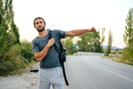 Young man hiker with backpack on hitchhiking near the road close up