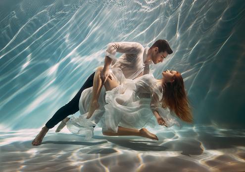 Real People art photo. Happy couple in love swim underwater, woman muse inspires male writer poet creator. Nymph girl dancing with guy at bottom sea under water. Red hair white long silk dress float.