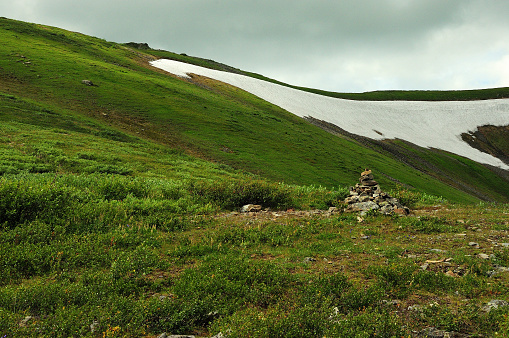Ritual stone pyramid on the gentle slope of a high mountain with grass and the remains of snow under a cloudy summer sky. Ivanovskie lakes, Khakassia, Siberia, Russia.