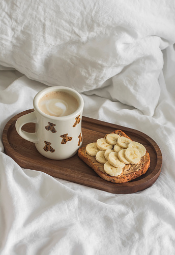 Toast with peanut paste and banana, cappuccino on a wooden tray on a bed with white linen bedding