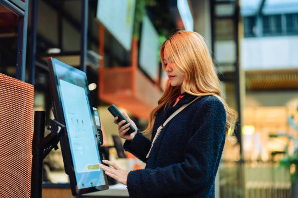 Woman at cashier counter uses smartphone for payment. Women in casual clothing paying using automatic payment machine and scanning through smartphone. contactless airport stock pictures, royalty-free photos & images