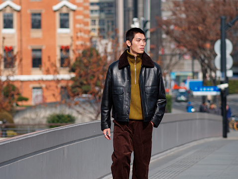 Portrait of handsome Chinese young man with black leather jacket posing with modern city street background in sunny winter day, male fashion, cool Asian young man.