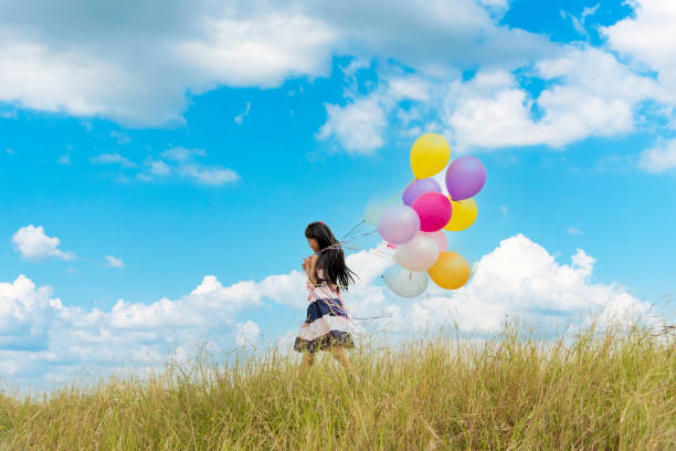 cheerful cute girl holding balloons running on green meadow white cloud and blue sky with happiness. hands holding vibrant air balloons play on birthday party happy times summer on sunlight outdoor - sun sky child balloon photos et images de collection
