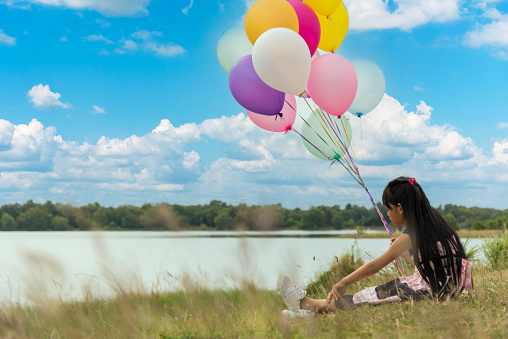 Cheerful cute girl holding balloons running on green meadow white cloud and blue sky with happiness. Hands holding vibrant air balloons play on birthday party happy times summer on sunlight outdoor
