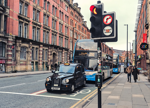 Manchester, United Kingdom - 12 29 2023 : A cab and a bus, stopped at a traffic light on a Manchester street. Perspective view of large brick wall in background.