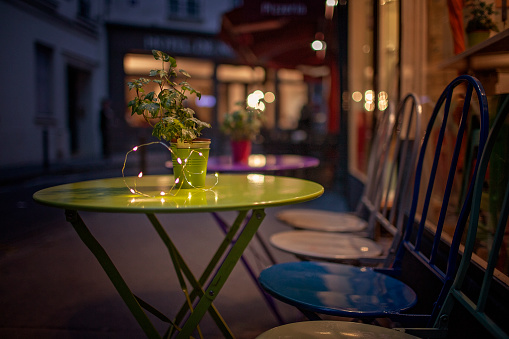 A cozy Parisian cafe terrace at dusk, with a green table hosting a potted plant and twinkling lights, offers an inviting atmosphere for evening relaxation and socializing. Travel and trip concepts