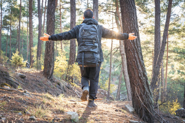 Young man opening his arms and embracing forest standing in between the trees with a backpack