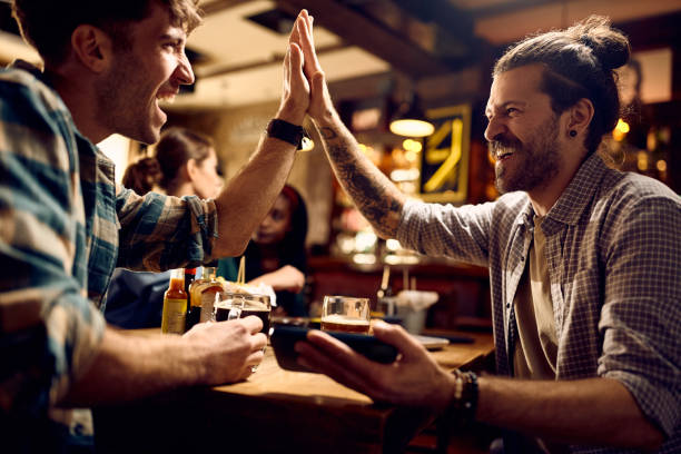 istock Cheerful men giving high-five while watching sports match on smart phone in a pub. 1936806384