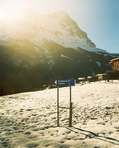 A Grindelwald Firstbahn sign in the snow with Swiss houses and a beautiful Mountain Vista in the background with stark morning light