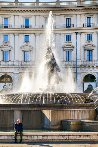 Rome, Italy, November 11 -- A man take a break near the beautiful Fontana delle Naiadi (Fountain of the Naiads) in Piazza della Repubblica, in the heart of Rome, on the edge of the Rione Monti. The Fountain of the Naiads was built in 1870 during the pontificate of Pope Pius IX, a few days before the conquest of Rome by the troops of the Kingdom of Italy and the end of the temporal power of the papacy, by the artists Mario Rutelli e Alessandro Guerrieri. In the photo: In the foreground the figure of a Nymph, while behind the figure of Glaucus grabbing a dolphin, symbol of man's primacy over nature. The Monti district is a popular and multi-ethnic quarter much loved by the younger generations and tourists for the presence of trendy pubs, fashion shops and restaurants, where you can find the true soul of the Eternal City. The Rione Monti, located between Via Nazionale and the Fori Imperiali, is also rich in numerous churches in Baroque style and archaeological remains. In 1980 the historic center of Rome was declared a World Heritage Site by Unesco. Image in high definition quality.