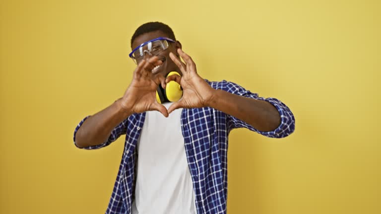 Handsome african american man in safety glasses standing against an isolated yellow background, showing love with symbolic heart shape hand gesture, radiating happiness in this romantic concept.