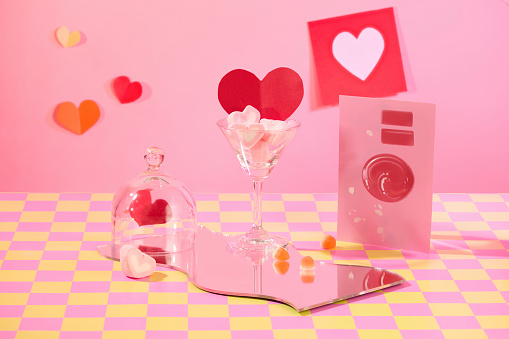 Heart-shaped marshmallow inside a cocktail glass, paper hearts, a card, an acrylic sheet and a glass dome cover. Pink-yellow checkered surface with pink background.