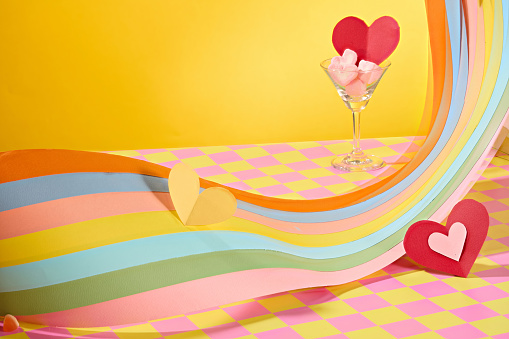 Heart-shaped marshmallows, a red paper heart in a cocktail glass, and a colored paper strip on display. Yellow background with a pink-yellow checkered surface. Copy space with front view.