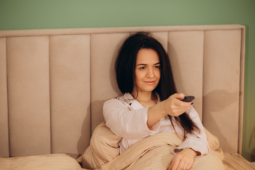 Woman watches TV sitting on the bed, using tv remote control in her hands. Morning or evening in the bedroom. Copy space. High quality photo