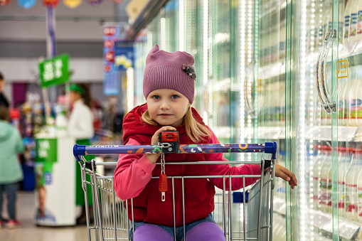 Little girl shopper 5 year old in red clothes get buy in supermarket, pensive looking at camera. Lovely kid girl sit in shopping cart buying in food store. Retail shopping concept. Copy ad text space