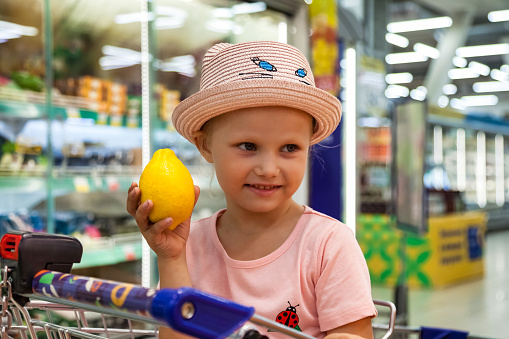 Happy child little girl sit in shopping cart holding lemon in hand in grocery store, smile looking away. Small kid girl in pink wear get buy in supermarket. Retail shopping concept. Copy ad text space