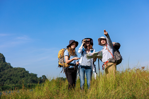 trekking family backpacker standing holding compass and map, while the other person pointing finger and using binoculars to look at view, blue sky backgroud,