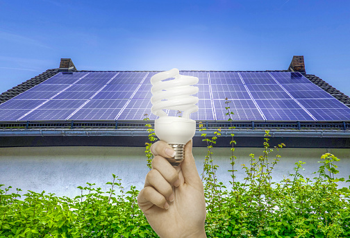 Hand holding a CFL bulb before solar panels on a house roof, with green foliage below, symbolizing sustainability. Energy saving concept