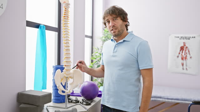 Bearded man in blue polo examining spine model at a bright rehabilitation clinic.